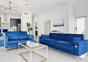A modern living room with two blue sofas, a glass coffee table, and a kitchen in the background. - Stunning Modern Oasis - Heated Pool & Spa · Mountain Views · 5min to Downtown! (Palm Springs)