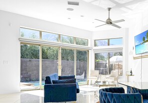A spacious living room with a large sliding glass door leading to a backyard, a ceiling fan, and a flat-screen TV mounted on the wall. - Stunning Modern Oasis - Heated Pool & Spa · Mountain Views · 5min to Downtown! (Palm Springs)