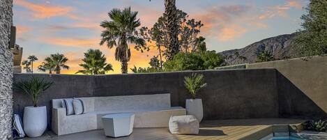 Un área de estar moderna al aire libre con piscina, rodeada por un muro de piedra y palmeras.