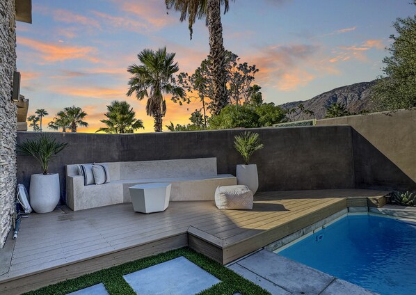 A modern outdoor seating area with a pool, surrounded by a stone wall and palm trees. - Stunning Modern Oasis - Heated Pool & Spa · Mountain Views · 5min to Downtown! (Palm Springs)
