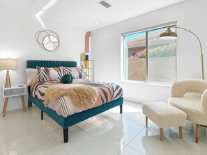 A modern bedroom with a teal bed, a white nightstand, a gold lamp, and a large window with a view of greenery. - Stunning Modern Oasis - Heated Pool & Spa · Mountain Views · 5min to Downtown! (Palm Springs)