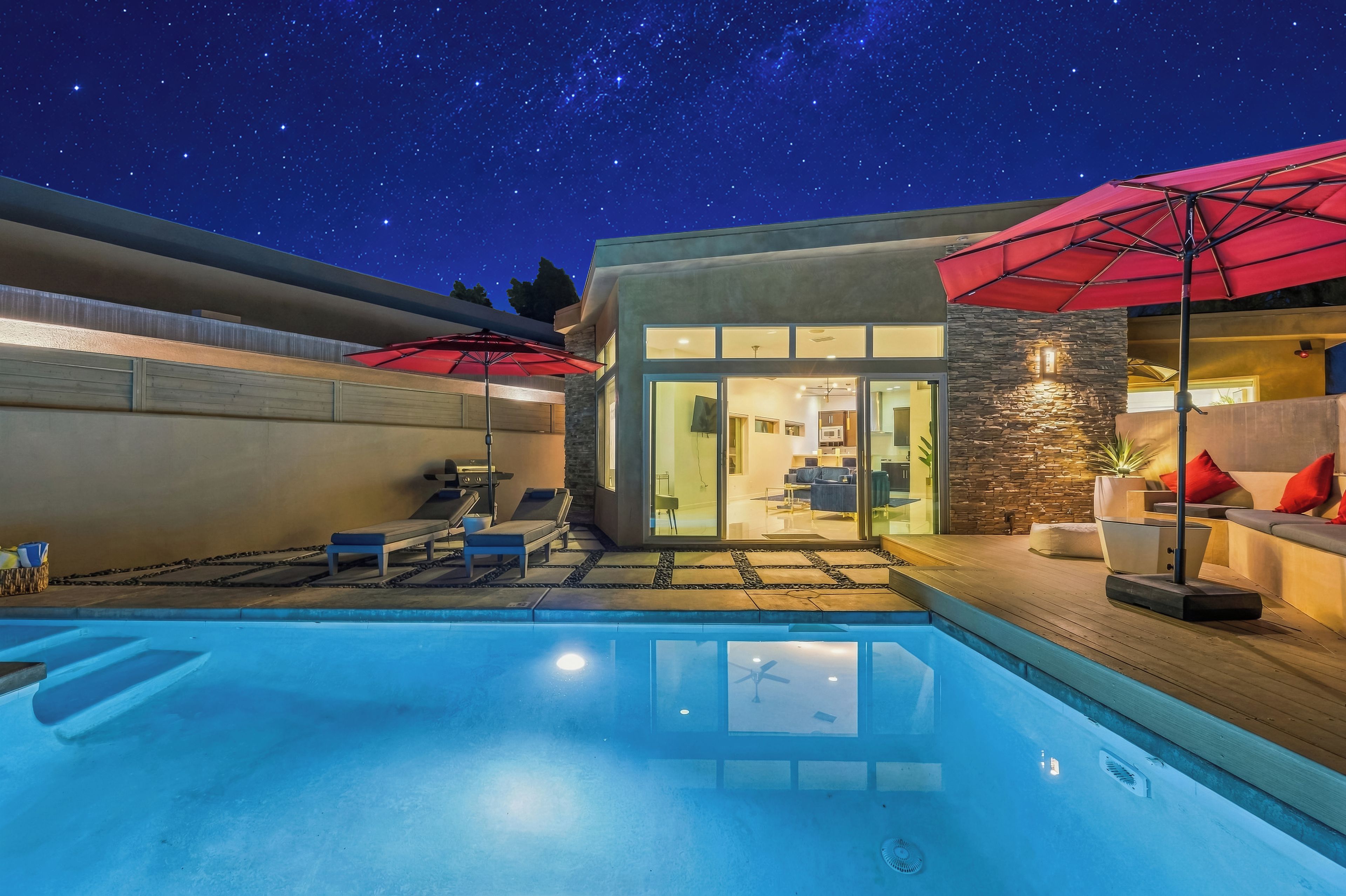 Un área de piscina exterior moderna con cielo nocturno despejado, pared de piedra y puerta de cristal que da a un espacio interior bien iluminado.