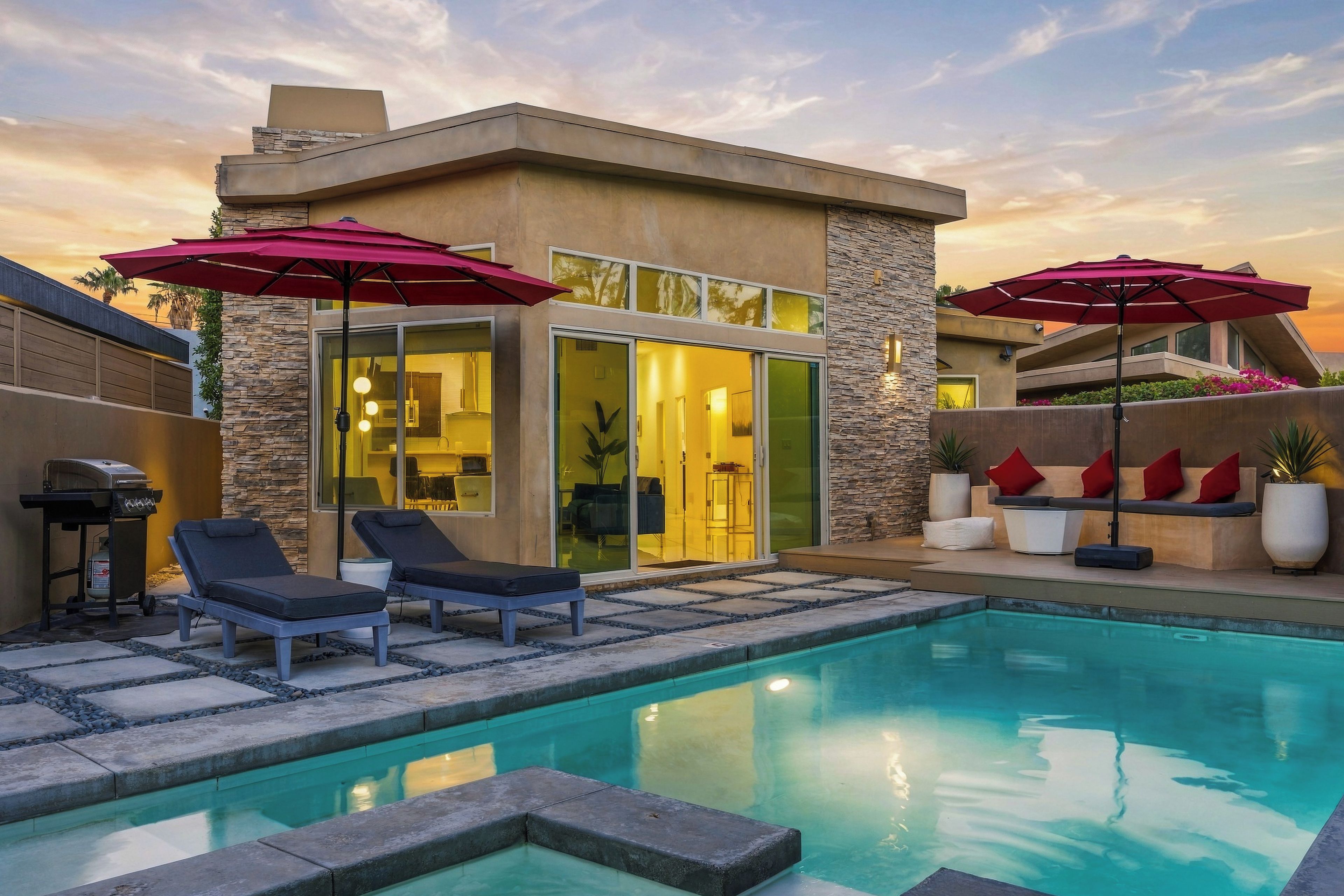 A modern outdoor pool area with lounge chairs and umbrellas, a stone wall, and a glass door leading to a bright interior space.