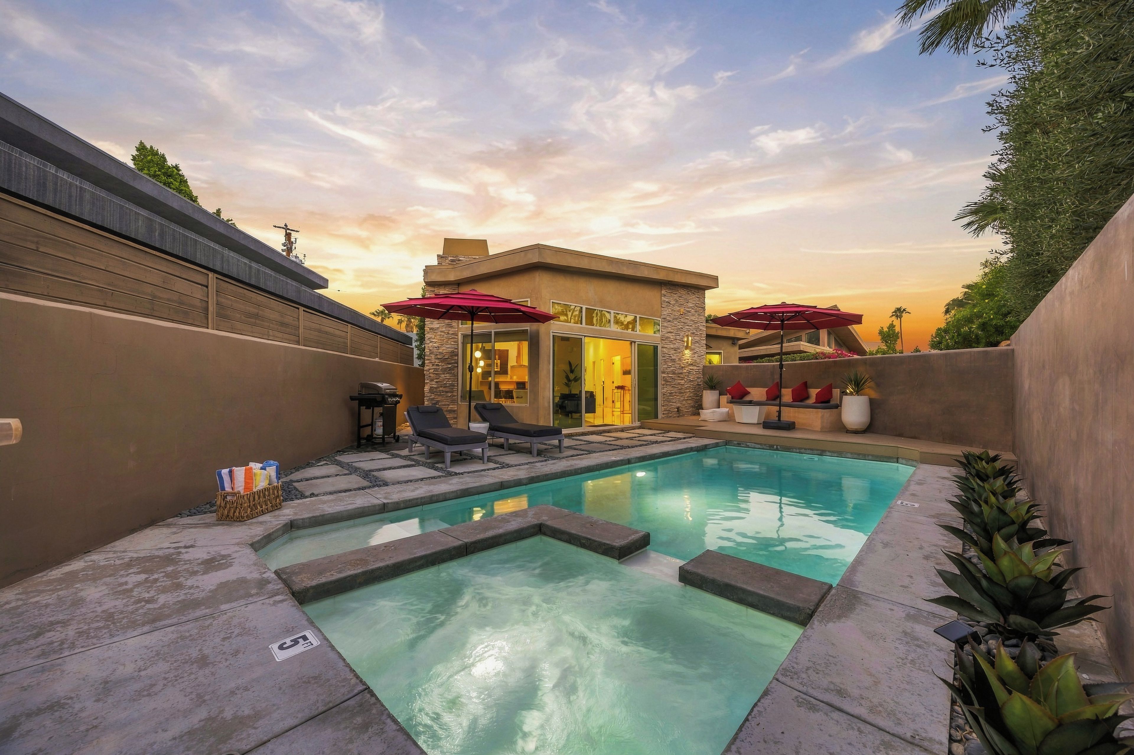A modern outdoor pool area with lounge chairs and umbrellas, surrounded by a stone wall and a building with a glass door.