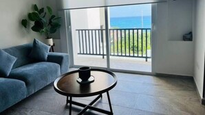Una habitación moderna con un sofá azul, una mesa de centro redonda y un gran ventanal corredero que da a un balcón con vistas al océano.