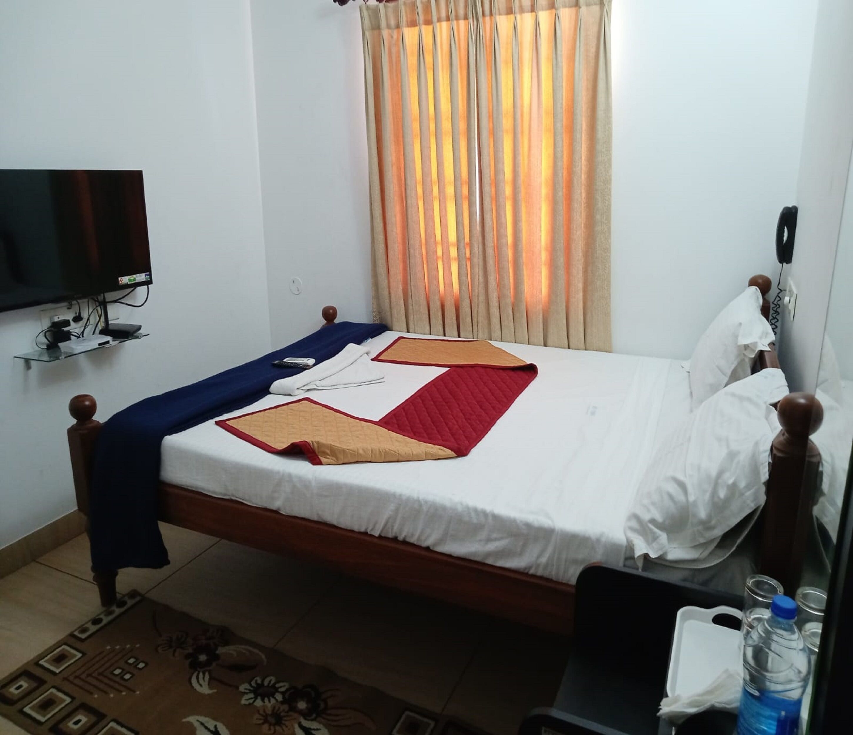 A single bed with a wooden frame, white bedding, and a red and beige quilt. A television is mounted on the wall to the left, and a telephone is on the right.
