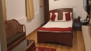 Une chambre avec un lit en bois, des coussins rouges, une chaise en osier, une petite table, un tableau au mur et un téléviseur sur le côté droit.