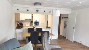 Una sala de estar moderna con un sofá gris, una mesa de centro de madera y un sillón gris. Al fondo, se ve una cocina con armarios blancos, una mesa de comedor con sillas y una lámpara colgante.