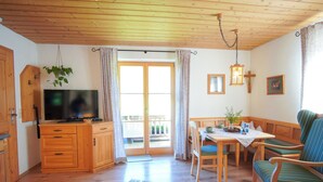 A wooden-floored living room with a television, wooden cabinets, a dining table with chairs, and a bench. - Ferienwohnung Veilchen für 2 Personen, 1 Separates Schlafzimmer, Balkon, 45m² (Inzell)
