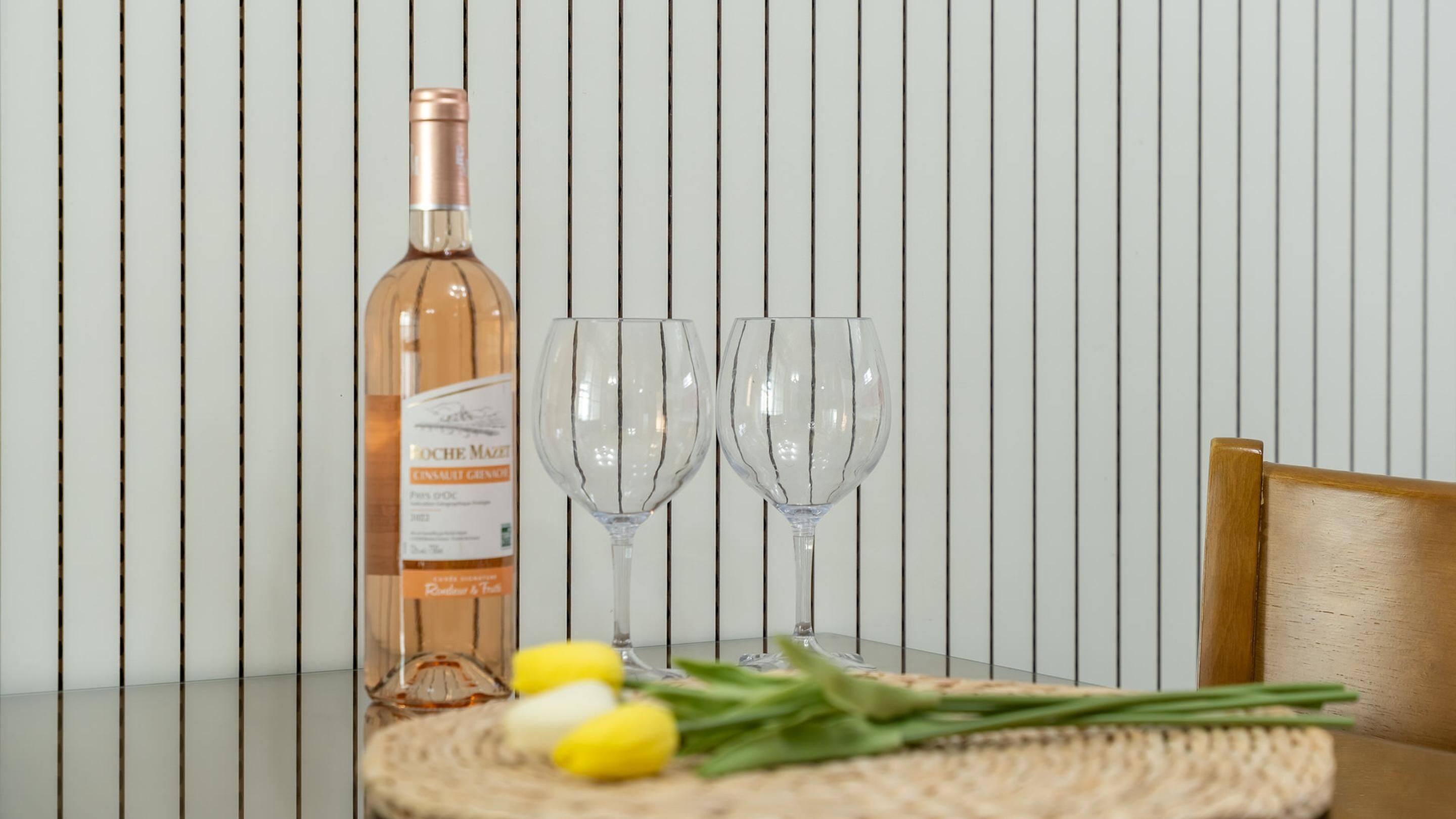 A bottle of wine, two wine glasses, and a woven placemat with yellow flowers on a table against a striped wall.