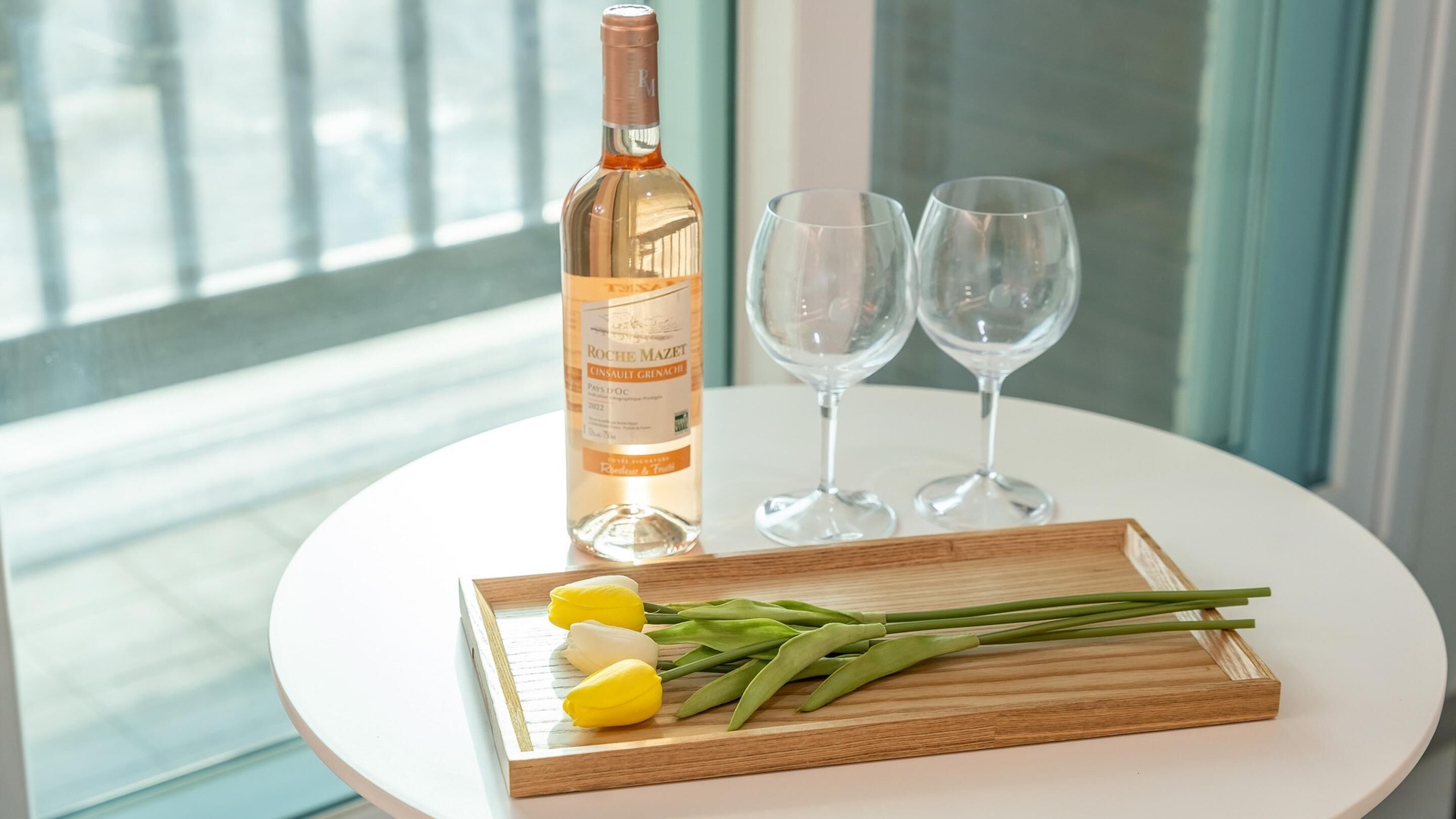 A bottle of wine, two glasses, and a wooden tray with vegetables on a white table by a window.