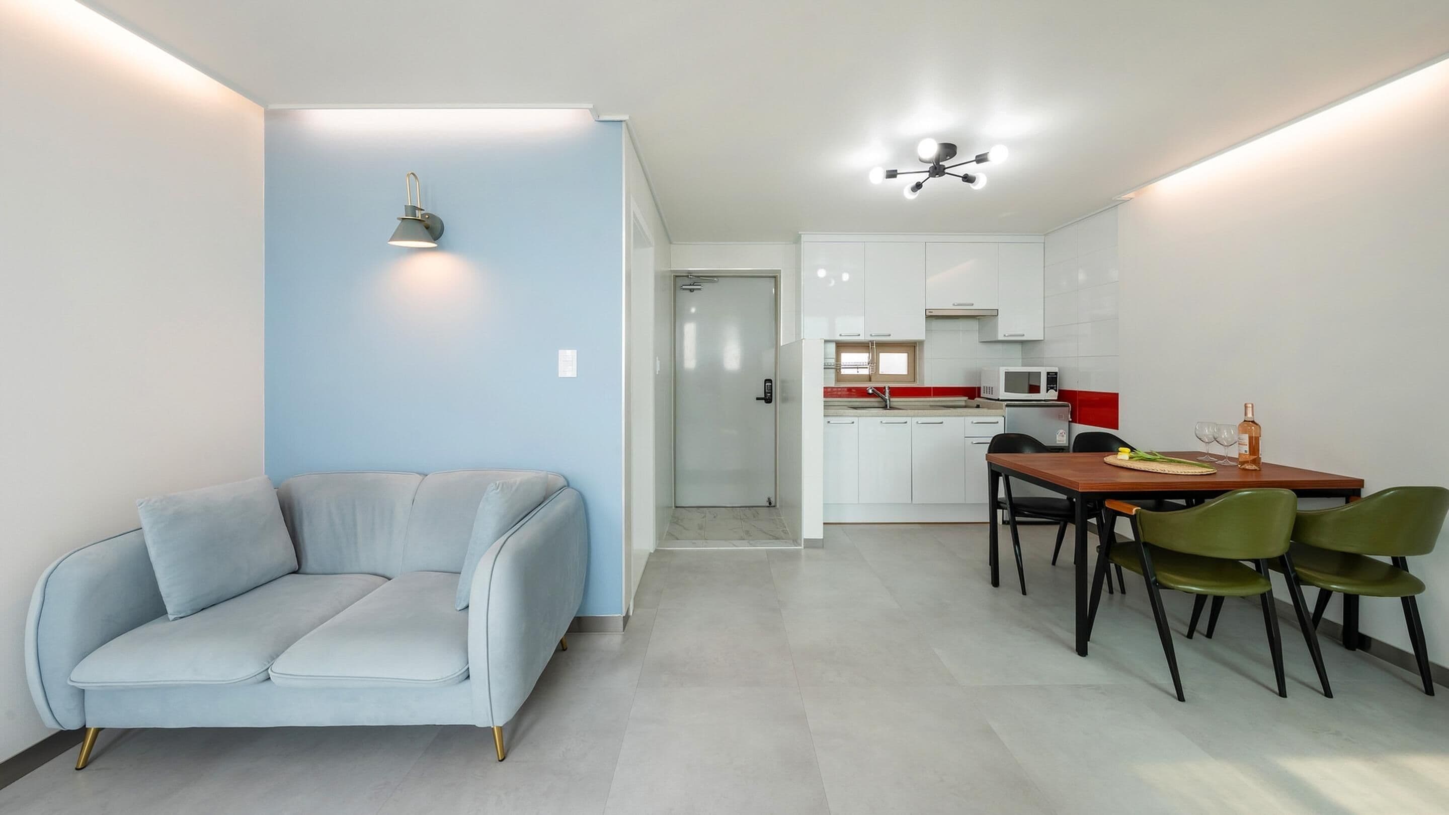 A modern living room with a light blue sofa, a dining area with a wooden table and green chairs, a kitchen with white cabinets, and a ceiling fan.