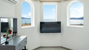 Een kamer met een grote flatscreen televisie aan de muur, een kaptafel met spiegel en een vaas rode rozen, en twee boogvensters met uitzicht op een wateroppervlak en verre bergen.