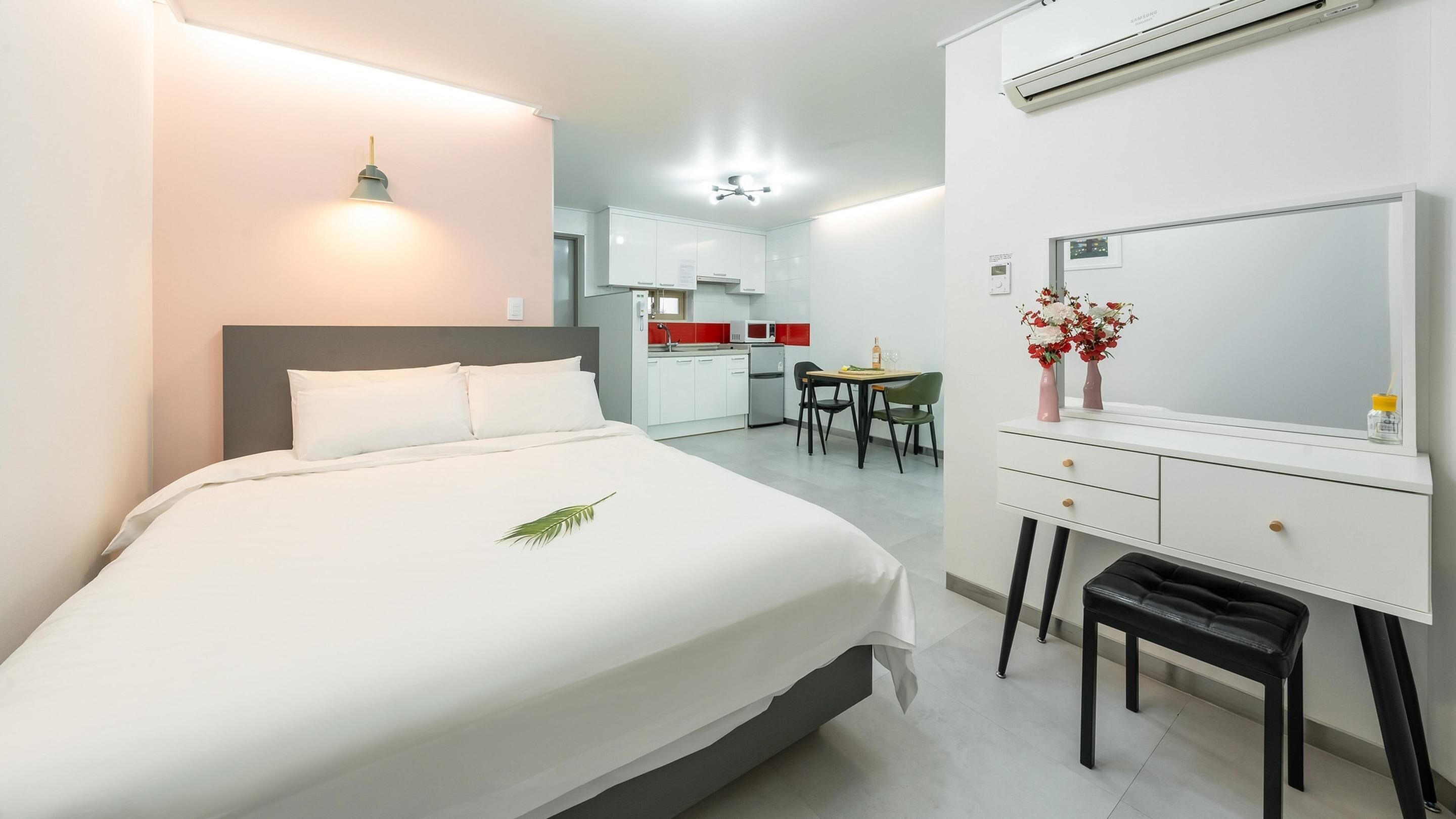 A hotel room with a bed, bedside table, and a desk with a chair. There is a kitchenette in the background.