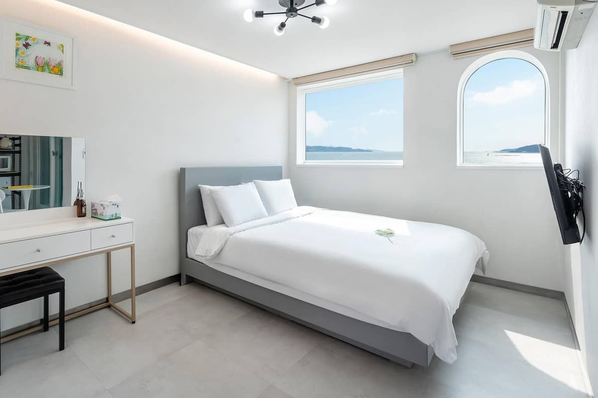 Ein modernes Schlafzimmer mit Bett, Schreibtisch, Stuhl, Spiegel und einem Fenster mit Meerblick.
