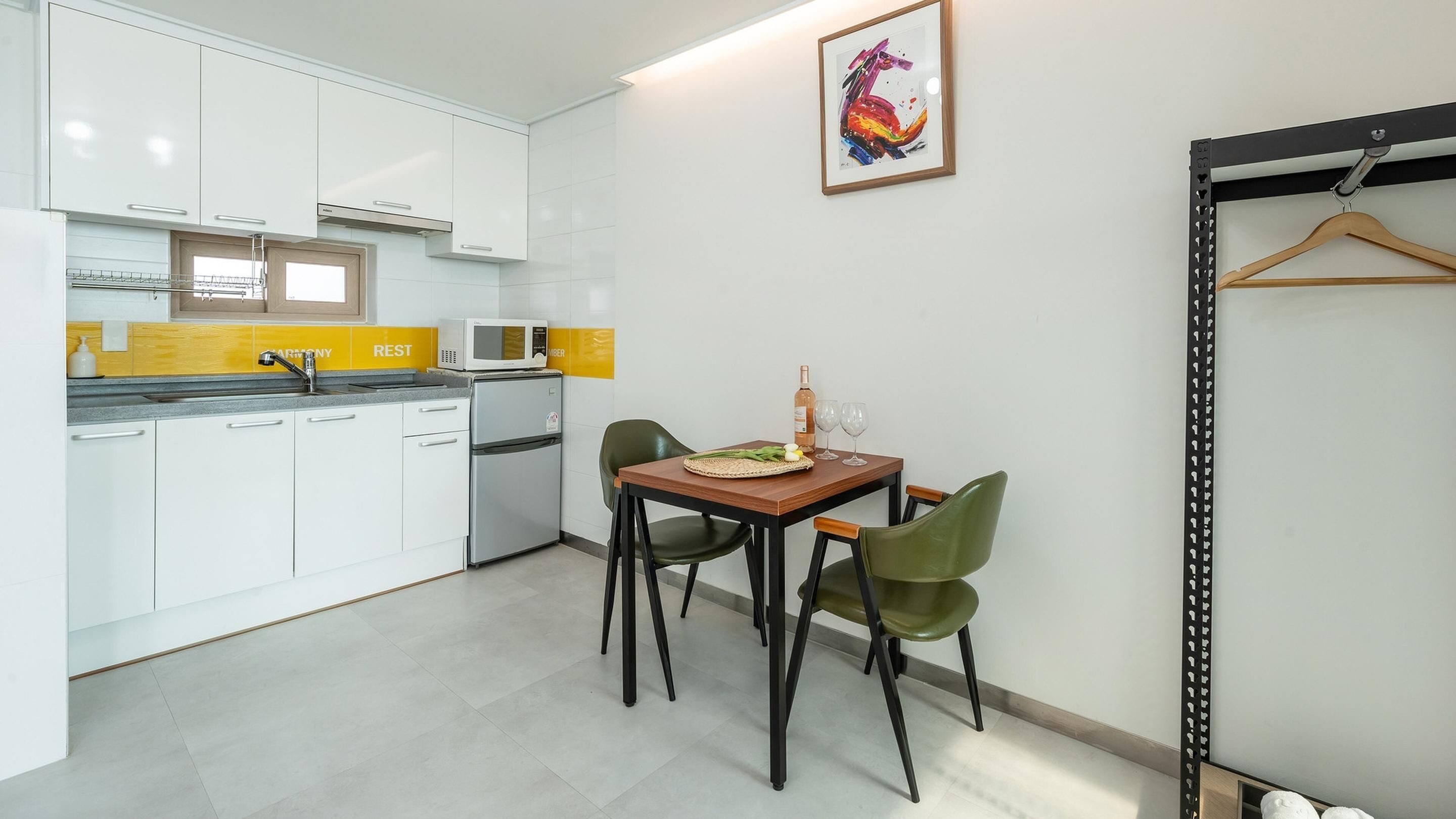 A modern kitchen with white cabinets, a microwave, and a small refrigerator. There is a dining table with chairs and a framed artwork on the wall.