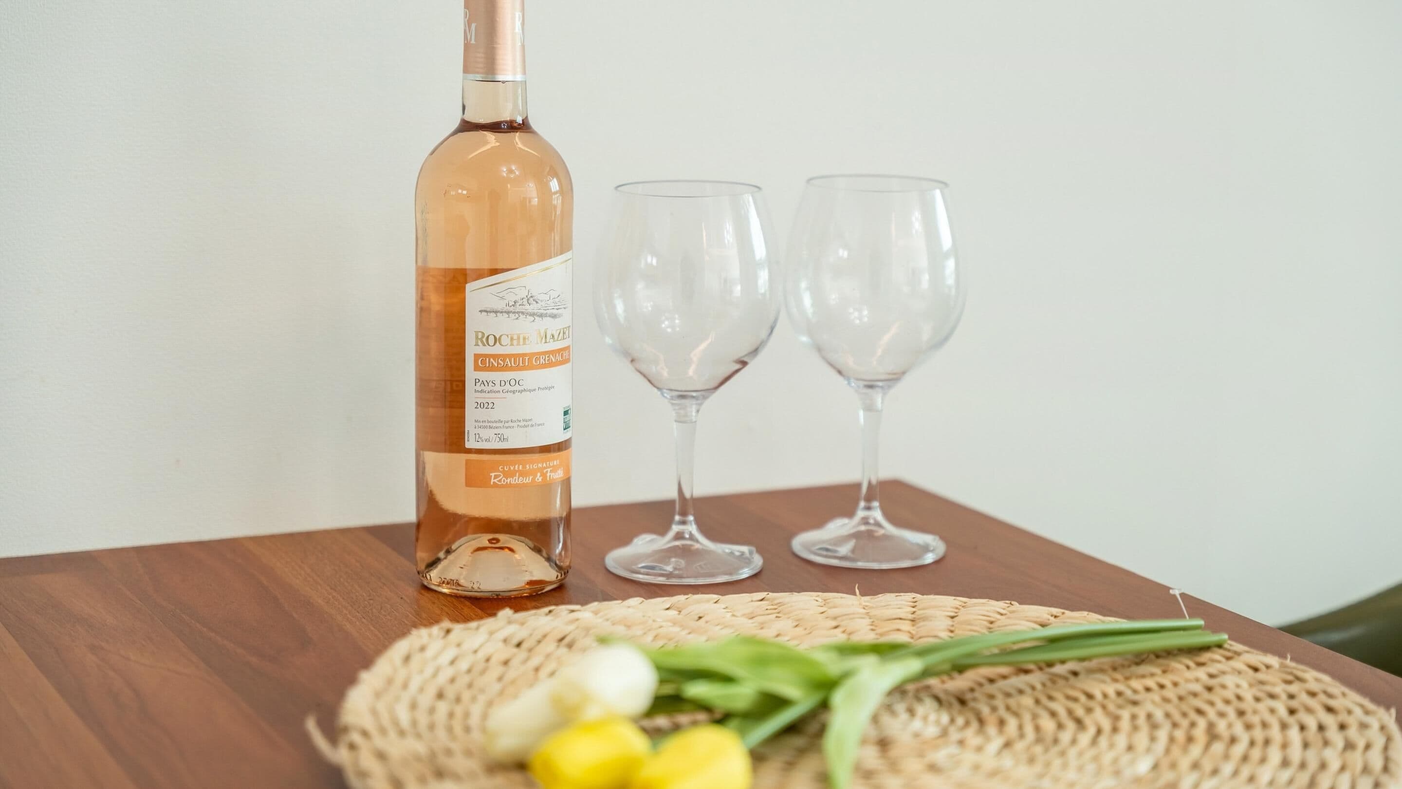 A bottle of wine, two empty wine glasses, and a woven placemat with some flowers on a wooden table.