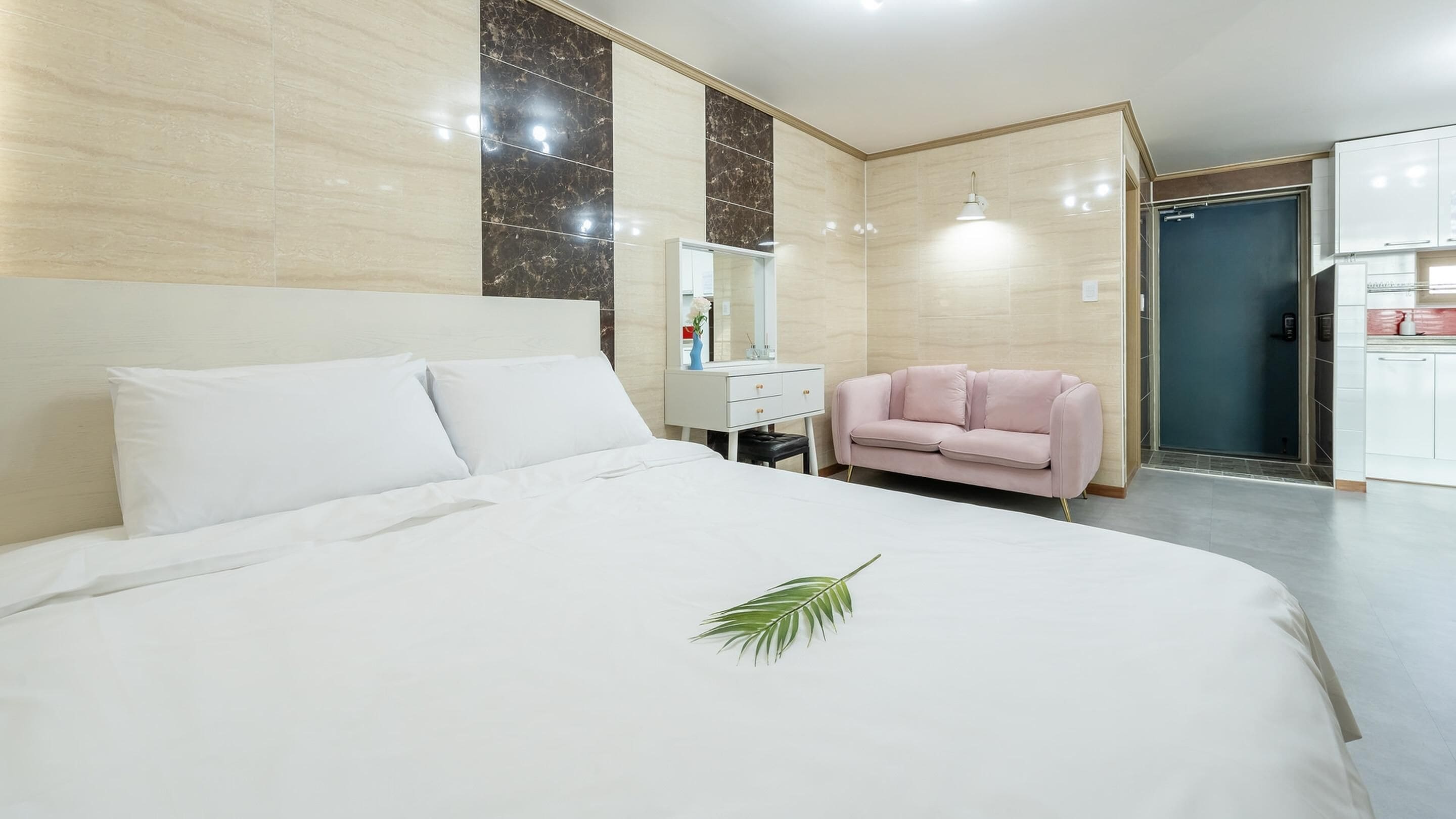 A hotel room with a large bed, a small desk, a pink chair, and a bathroom visible through an open door.
