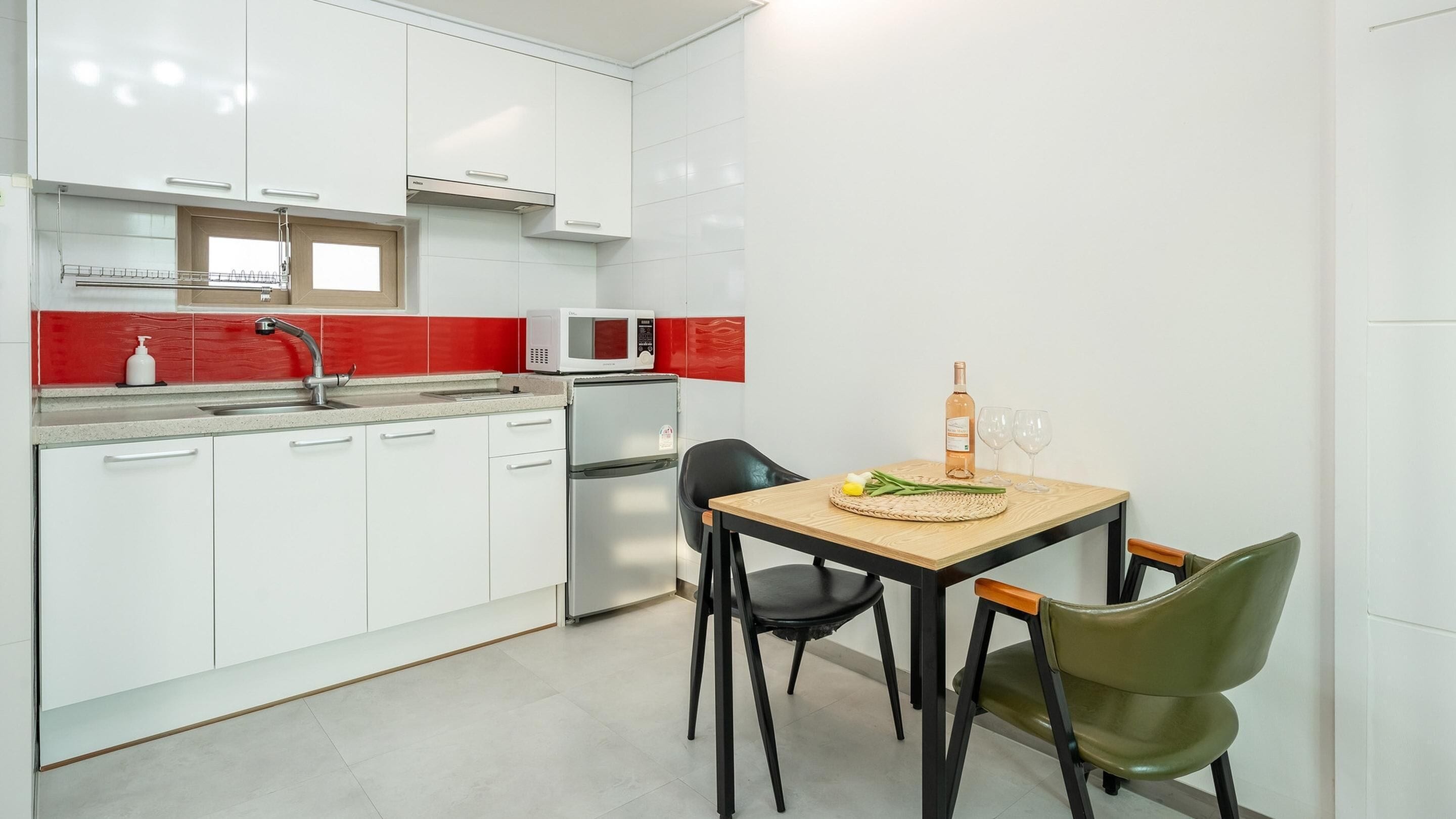 A modern kitchen with white cabinets, a red backsplash, a microwave, a refrigerator, and a dining area with a table and chairs.