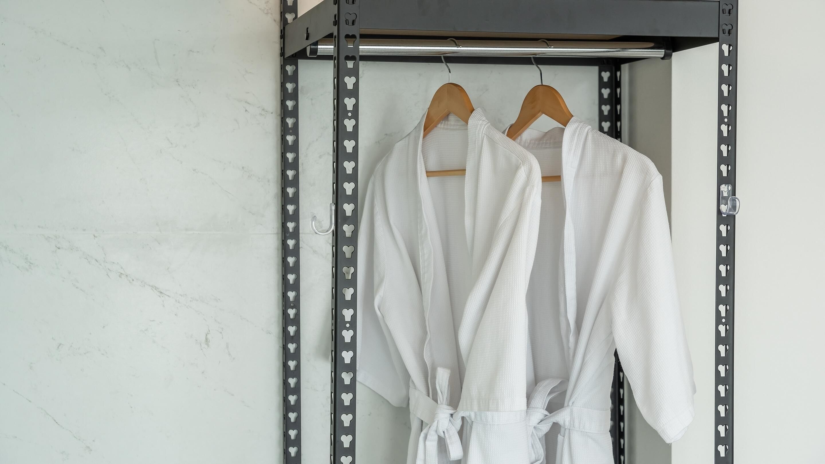 A bathroom with a marble wall, a metal rack with heart-shaped cutouts, and two white bathrobes hanging on wooden hangers.
