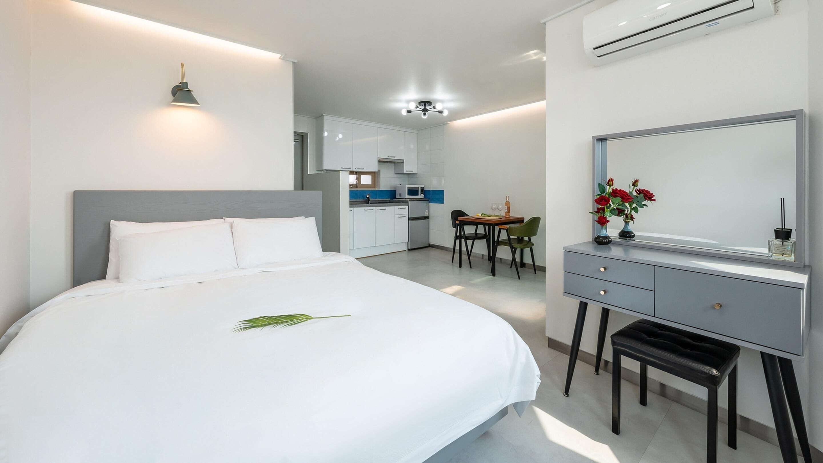 A modern hotel room with a large bed, a desk, a chair, and a kitchenette in the background.