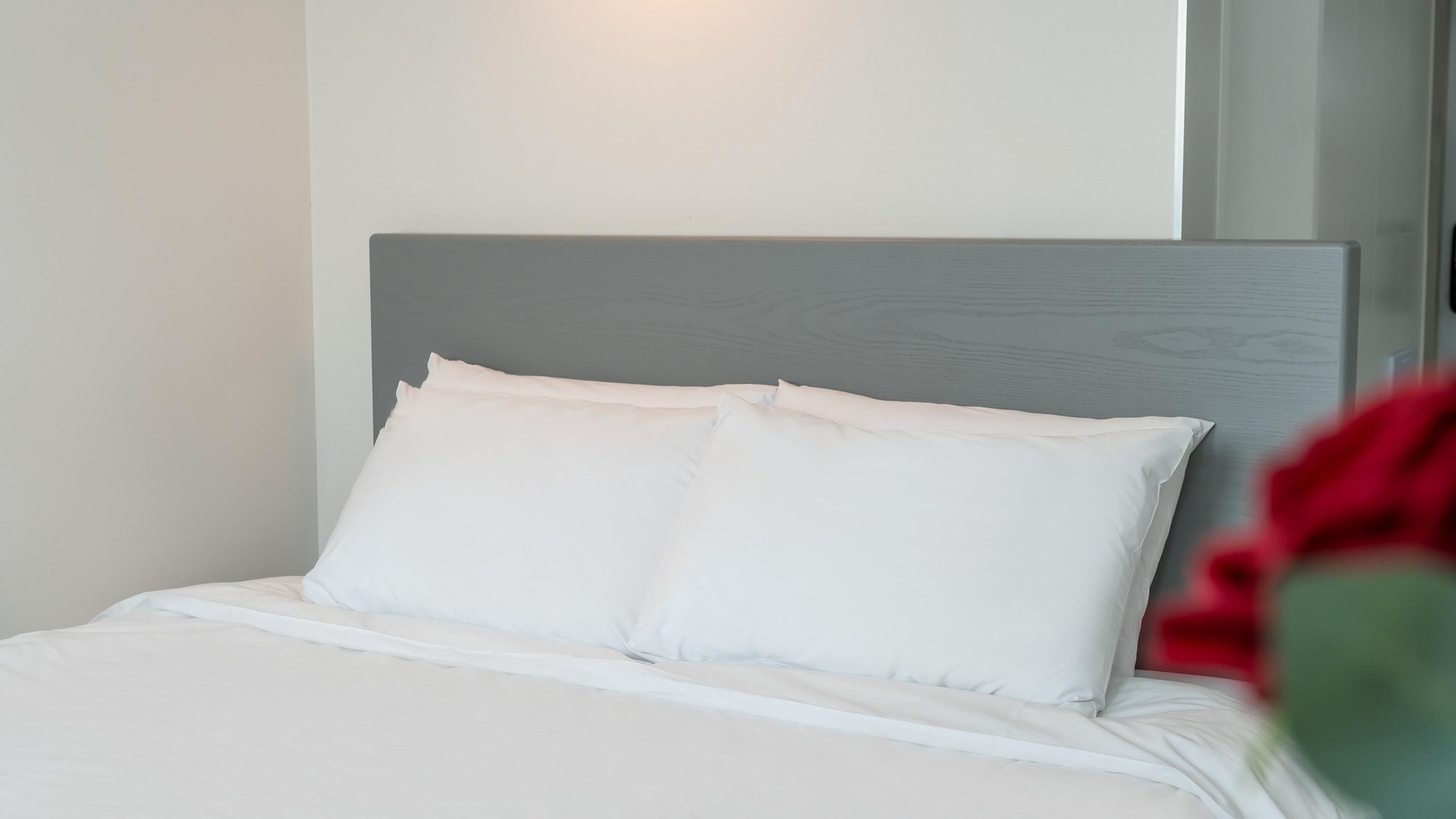 Una cama bien hecha con ropa blanca y almohadas, cabecera gris y una lámpara fijada a la pared.