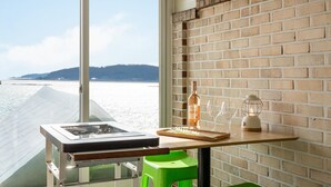 Een kamer met een groot raam dat uitkijkt op een strand, een bakstenen muur, een tafel met een fles, glazen en een lantaarn.