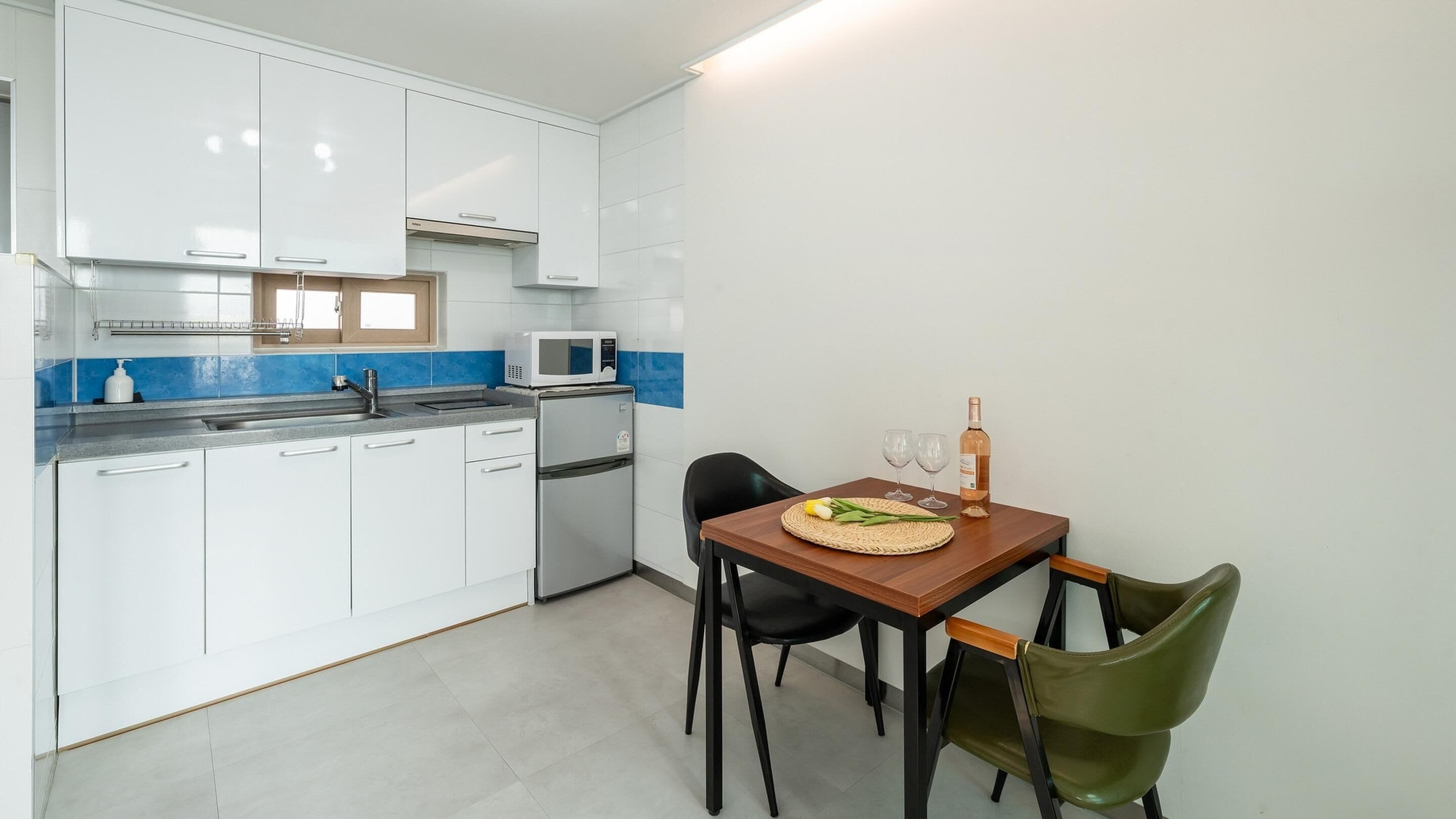 A compact kitchen with white cabinets, a microwave, and a small refrigerator. There is a dining table with two chairs and a bottle of wine.