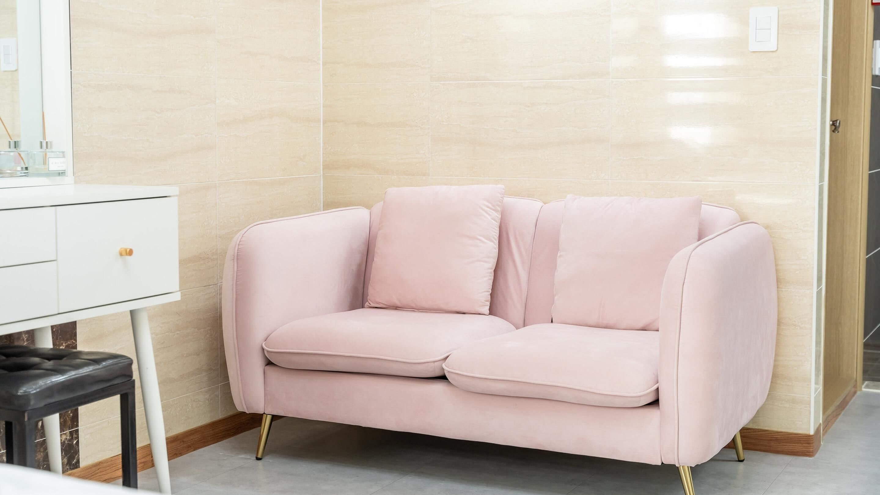 Un sofá rosa con patas doradas en una habitación con paredes de baldosas color beige y un armario blanco.