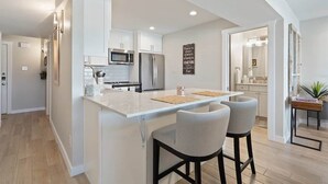 A modern kitchen with white cabinetry, a central island with bar stools, a microwave, and a refrigerator. - Luxury Waterfront Condo  Pool + Hot tub  Sleeps 4 (Spicewood)