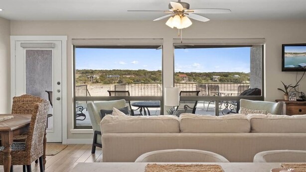 A room with a ceiling fan, a sofa, a dining table with chairs, and a view of a beach and houses through large windows. - Luxury Waterfront Condo  Pool + Hot tub  Sleeps 4 (Spicewood)