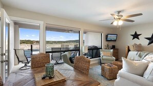 A spacious living area with a large window overlooking a scenic view, a ceiling fan, and a comfortable seating arrangement. - Luxury Waterfront Condo  Pool + Hot tub  Sleeps 4 (Spicewood)
