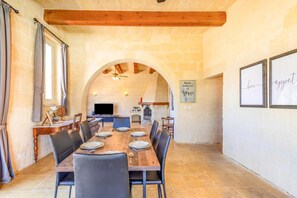 A dining area with a long wooden table, black chairs, and a stone wall with an arched doorway leading to a kitchen. - Nirvana Gozitan Villa & Pool (Qala)