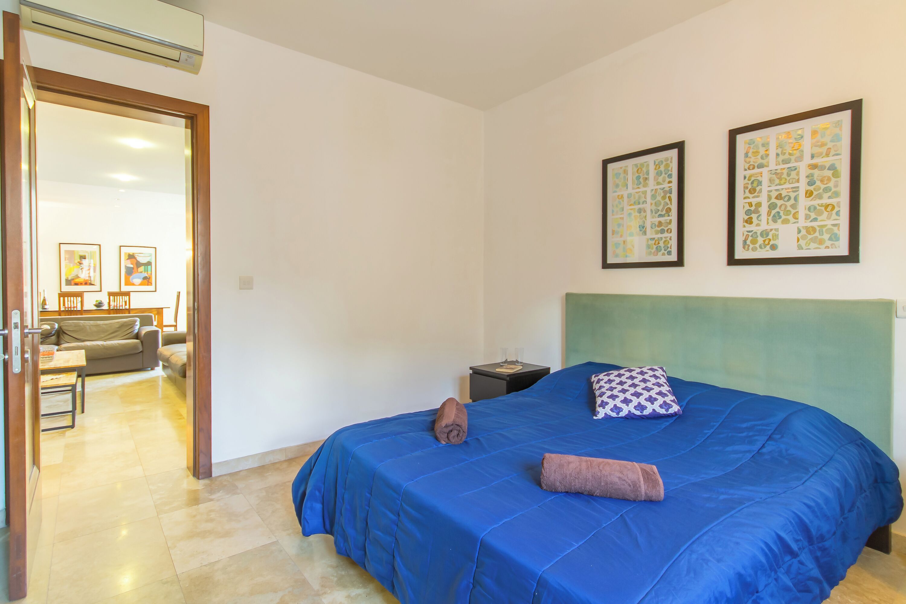 A bedroom with a blue bedspread, a green headboard, and two framed artworks on the wall.