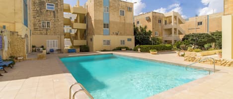 Una piscina rodeada de edificios de varios pisos con balcones, un muro de piedra y plantas en macetas.