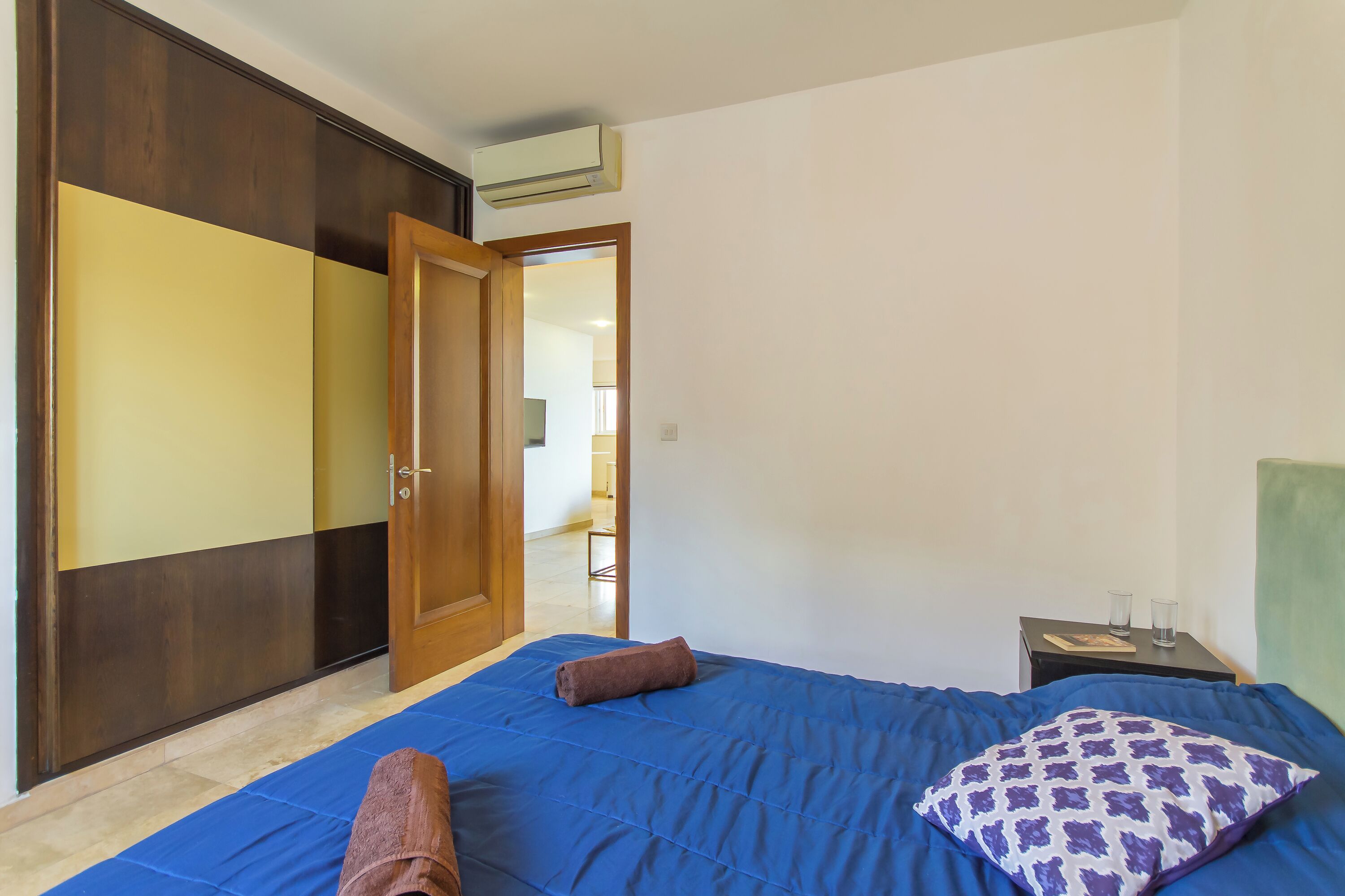 A bedroom with a bed, a wooden wardrobe, a nightstand, and an open door leading to another room.