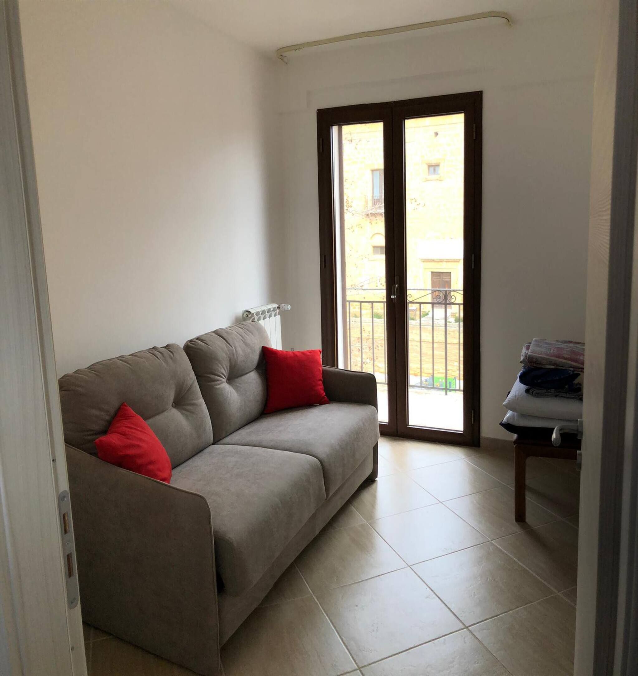 A living room with a sofa, red cushions, a radiator, a sliding glass door to a balcony, and a small table with folded items.