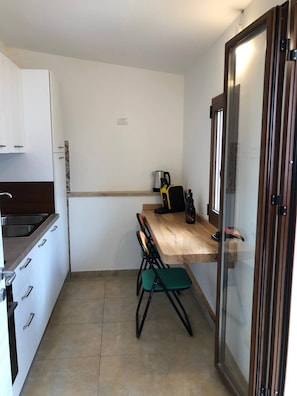 Cocina compacta con alacenas blancas, encimera de madera, un lavabo y una zona de comedor con una silla.