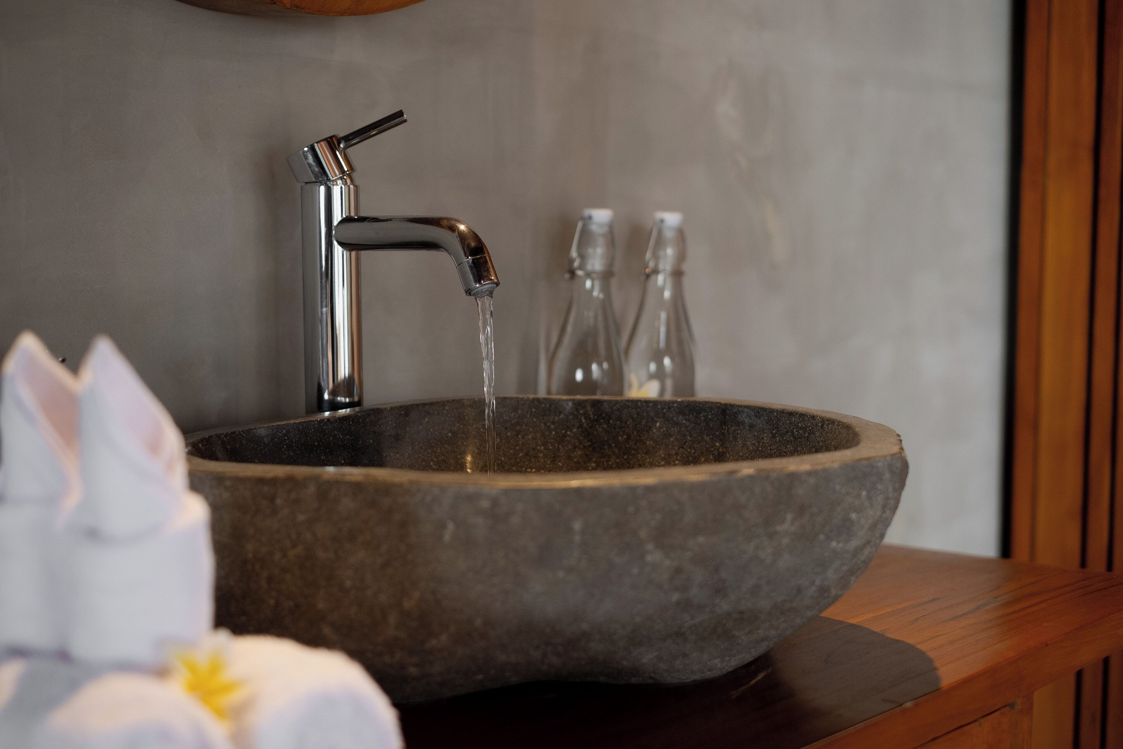 Ein Waschbecken aus Stein mit einem modernen Wasserhahn, eine Holzauskastung und zwei klare Glasflaschen im Hintergrund.