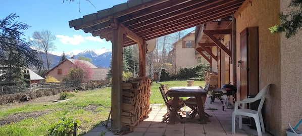 Une terrasse couverte avec une table et des chaises, un barbecue et une vue sur les montagnes en arrière-plan.