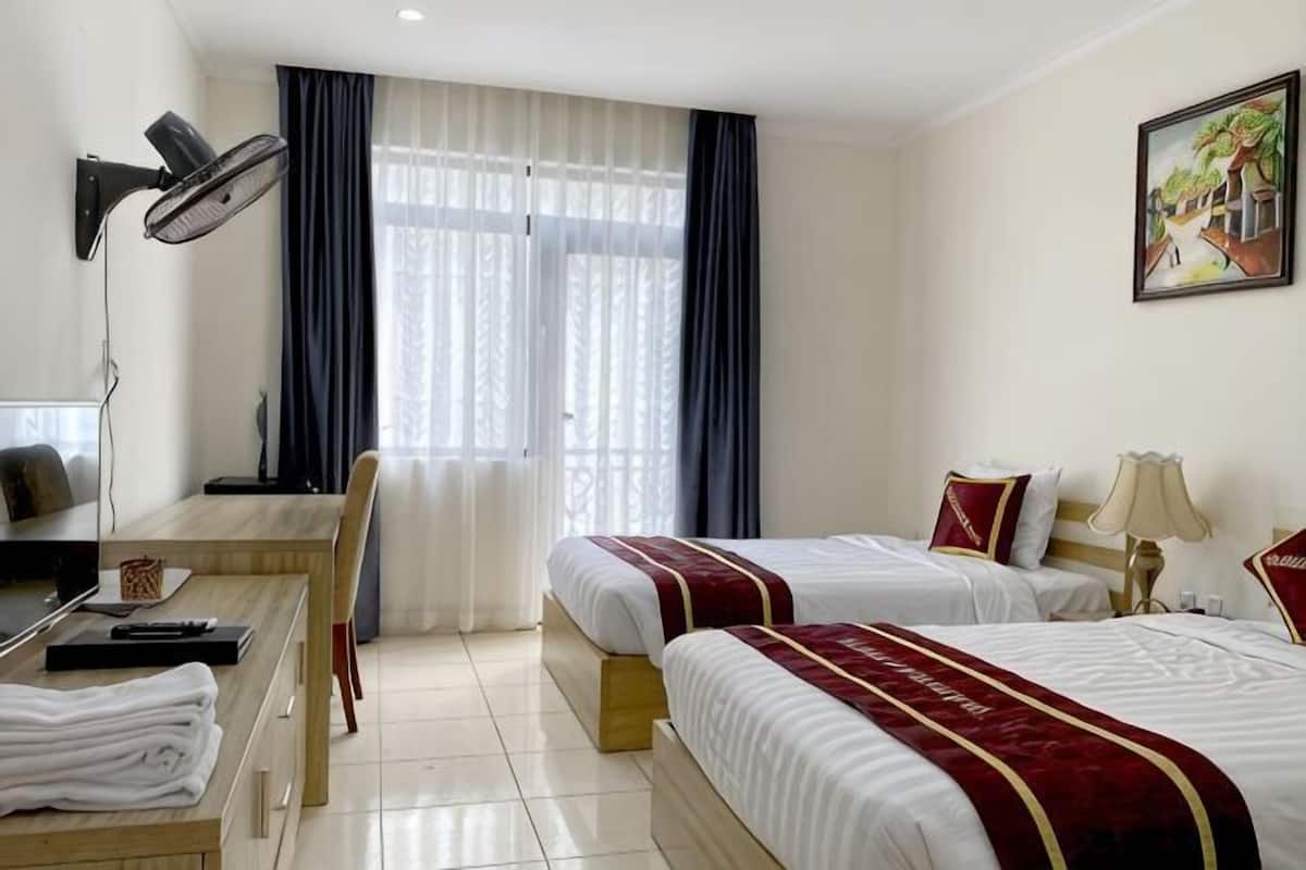 A hotel room with two beds, a desk, a chair, a desk lamp, a window with curtains, and a painting on the wall.