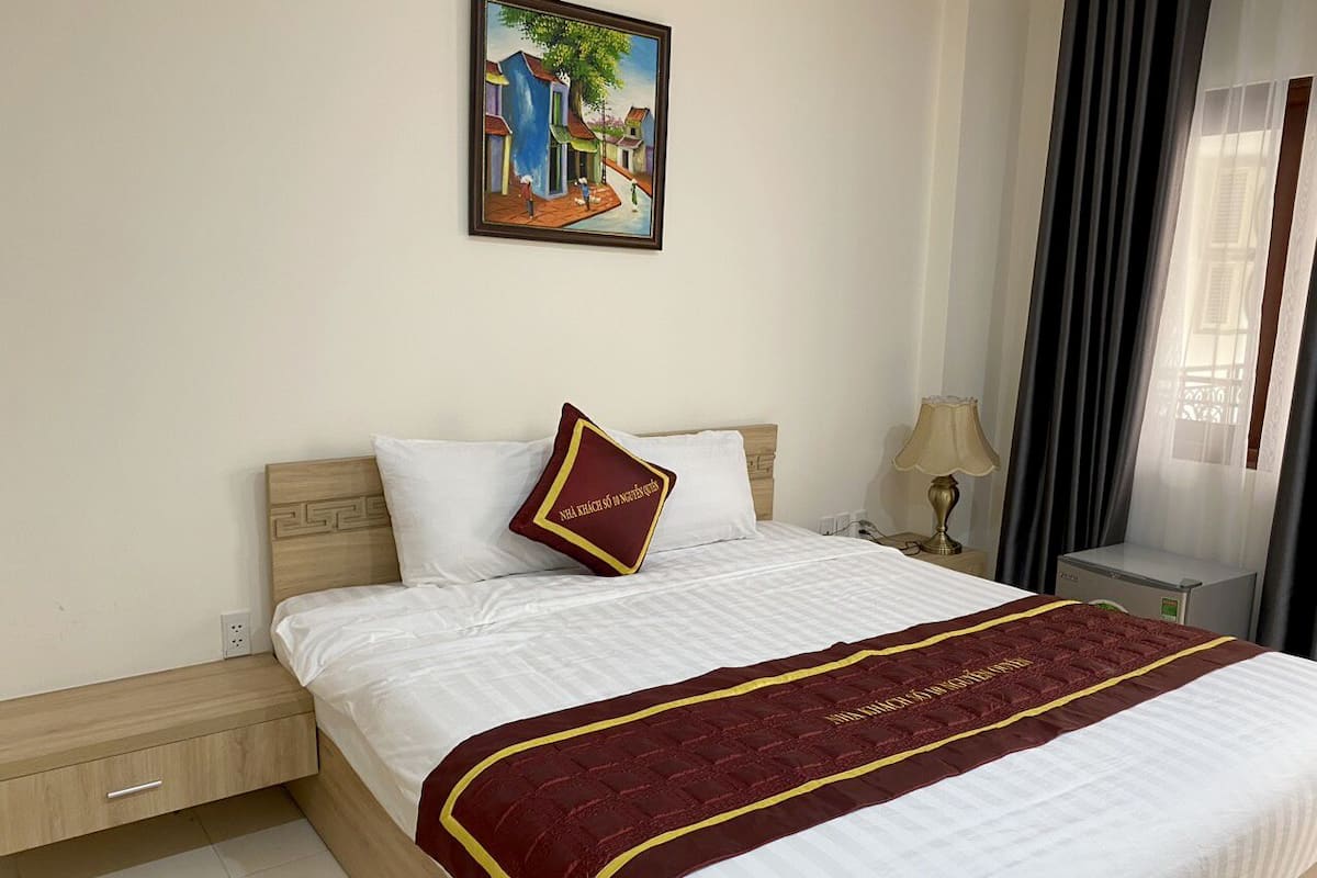 A hotel room with a bed, a nightstand, a lamp, a painting on the wall, and a window with curtains.
