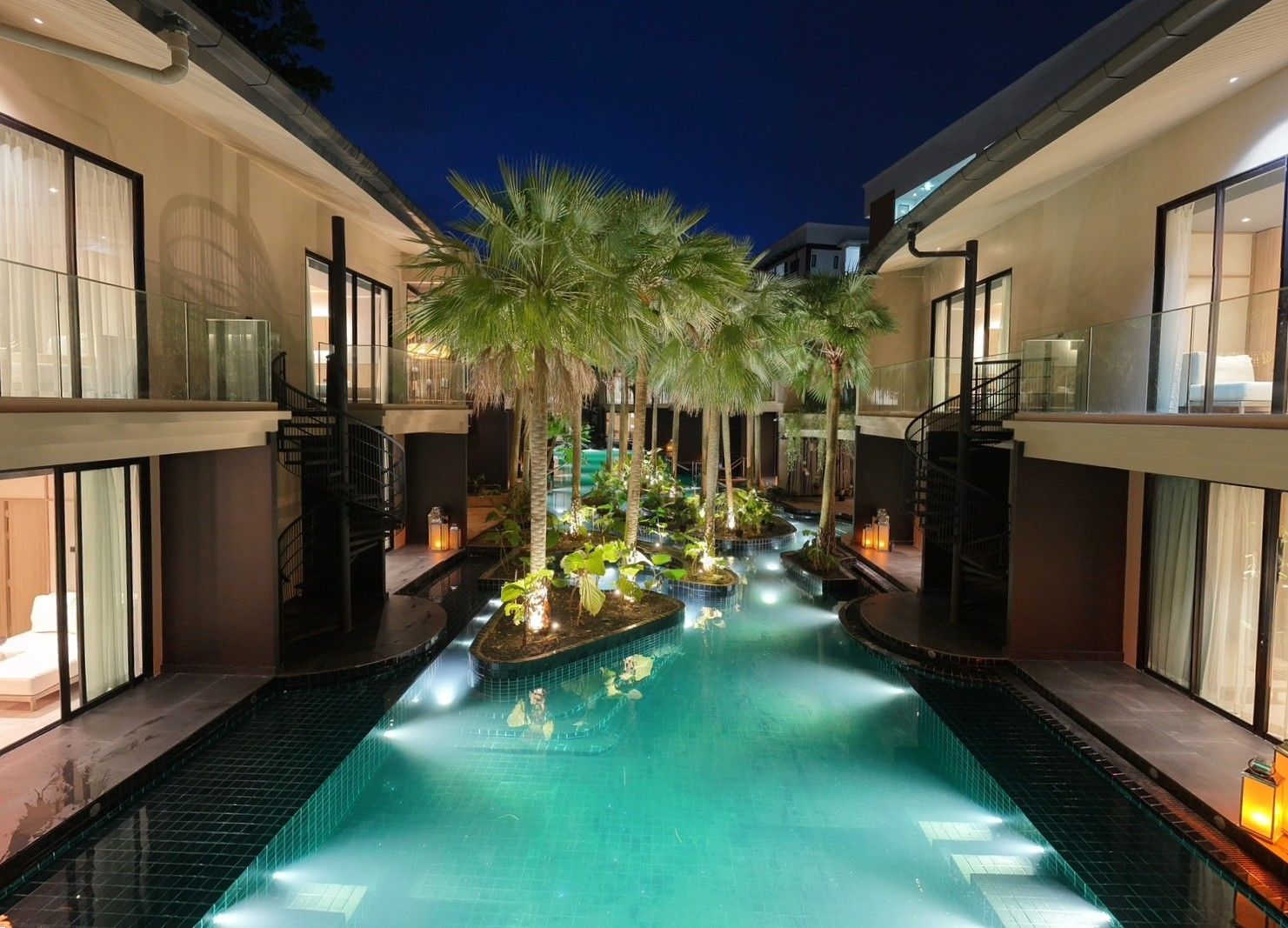A modern hotel with a central swimming pool, surrounded by palm trees and illuminated by ambient lighting.