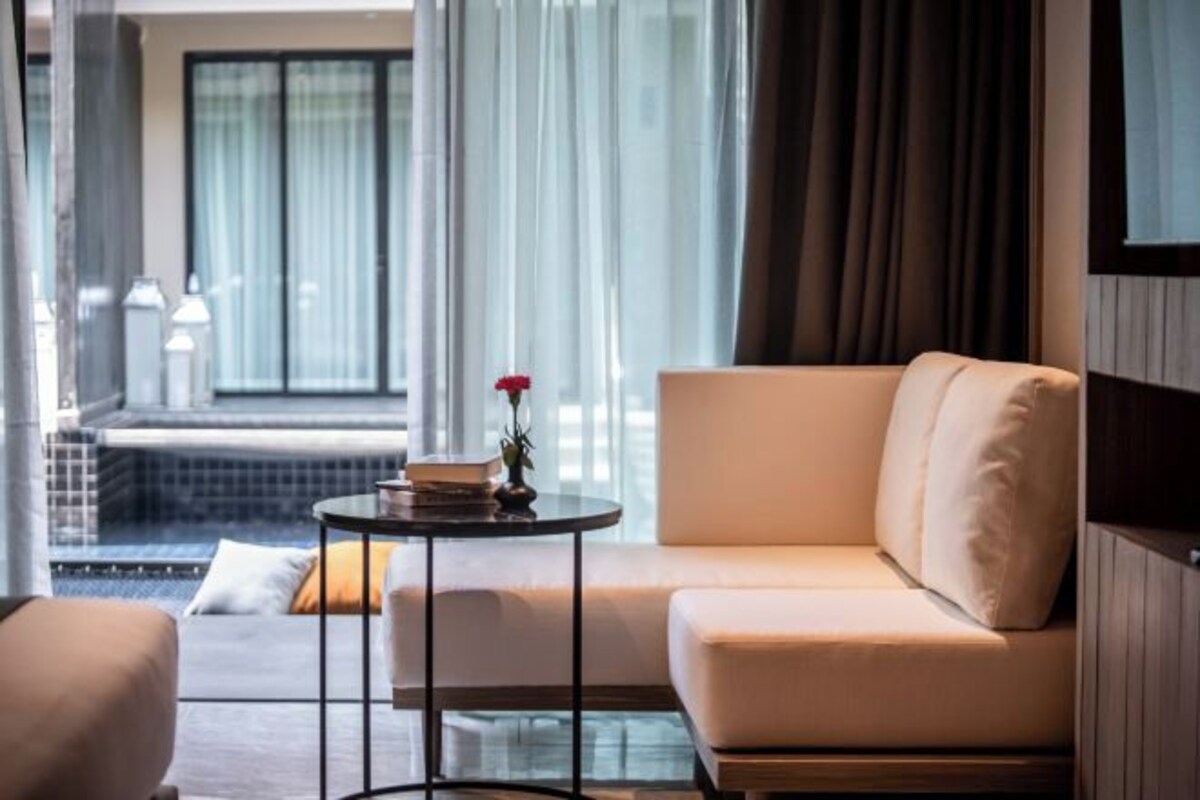 A modern hotel room with a sofa, a small round table with a book and a rose, and a view of a pool and outdoor area through large windows.