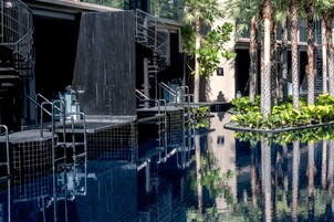 Piscina con escalones de cerámica y fuente de agua, rodeada de palmeras y vegetación.