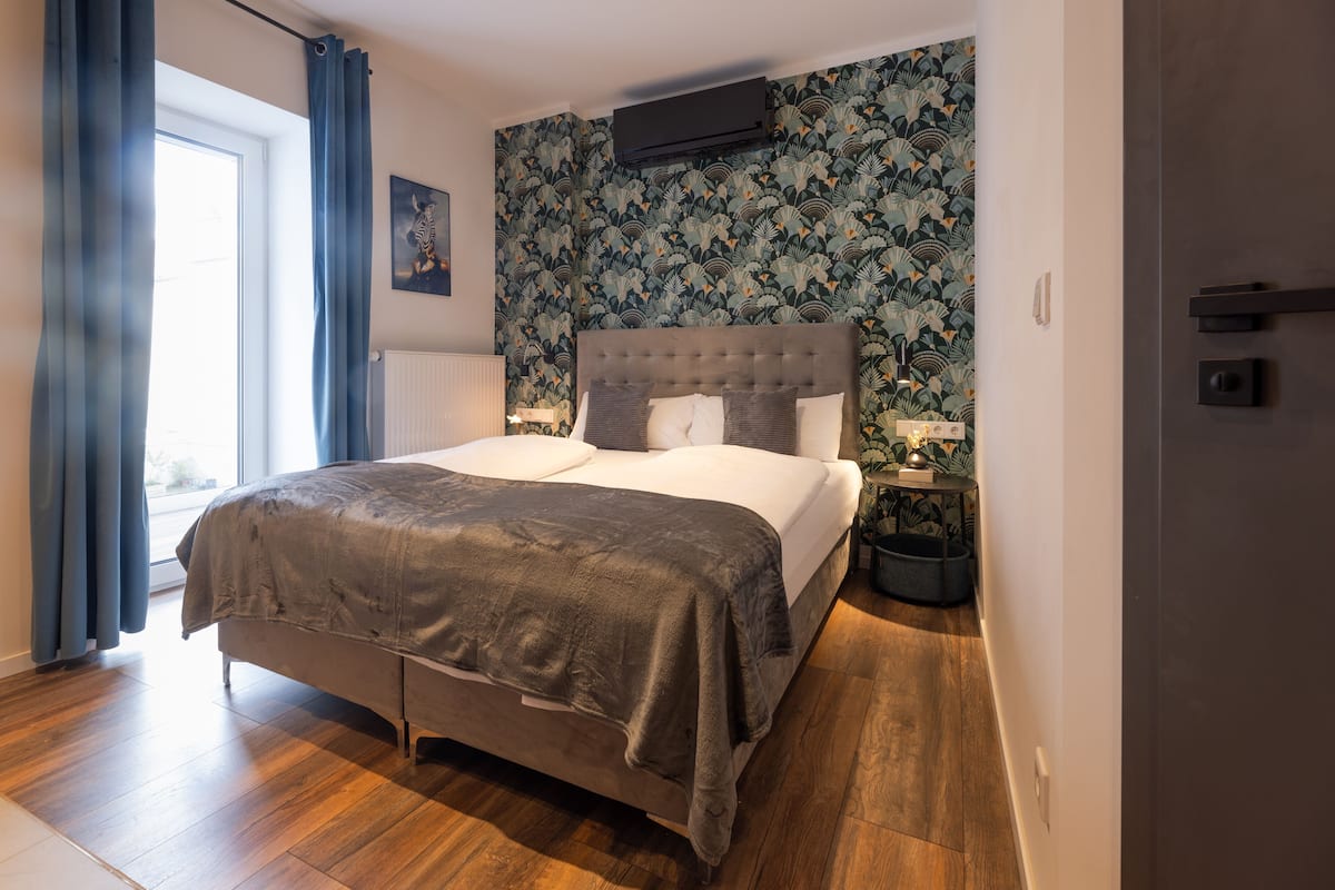 A modern bedroom with a large bed, a wall-mounted air conditioner, and a patterned wallpaper.