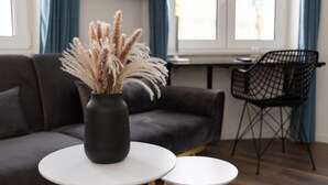 Una sala de estar moderna con un sofá gris oscuro, una mesa redonda blanca con un jarrón de plumas de pluma y una silla de alambre negra.