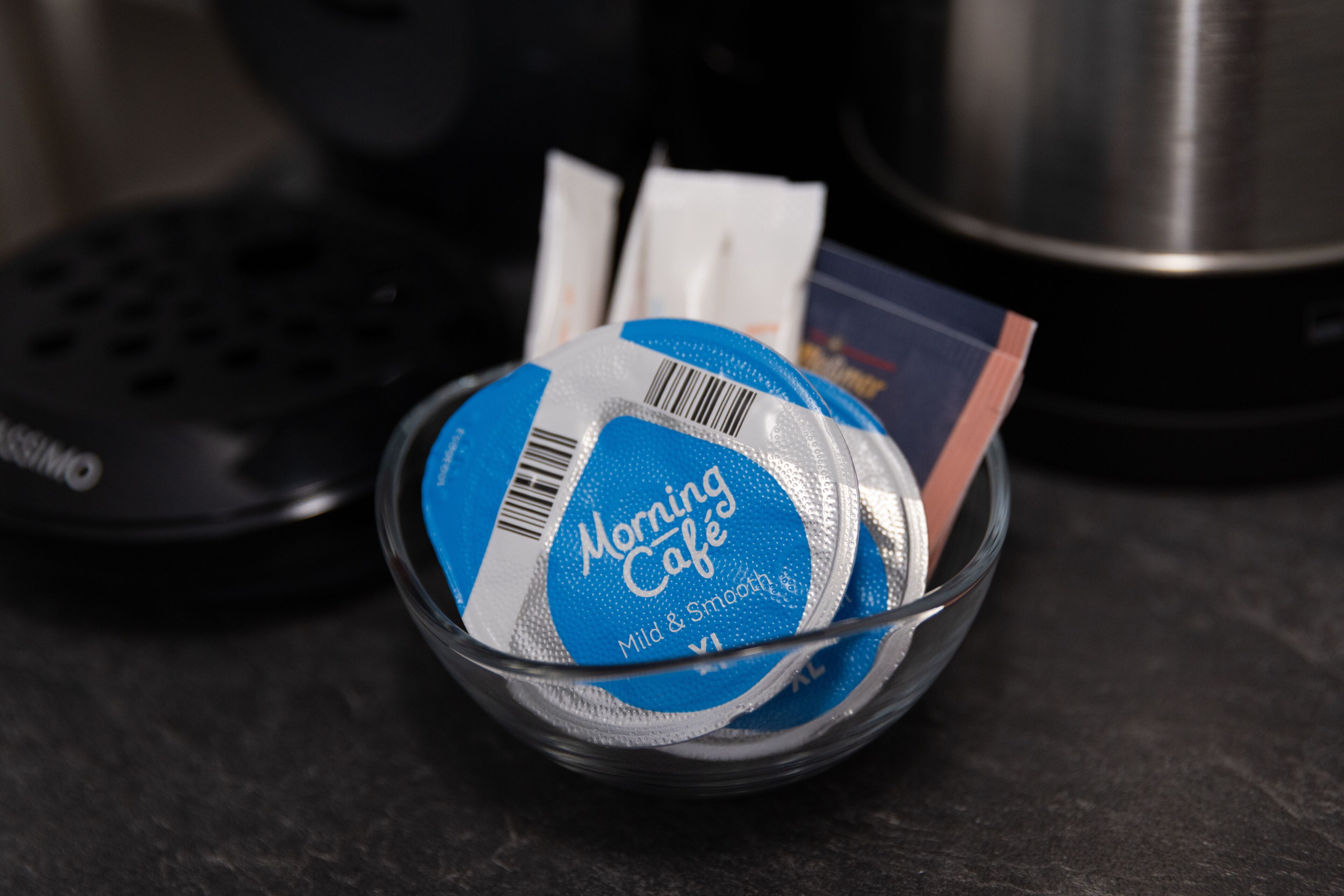 En glasskål som innehåller ett blått paket märkt med texten ”Morning Café” och en streckkod, placerad på en mörk yta.