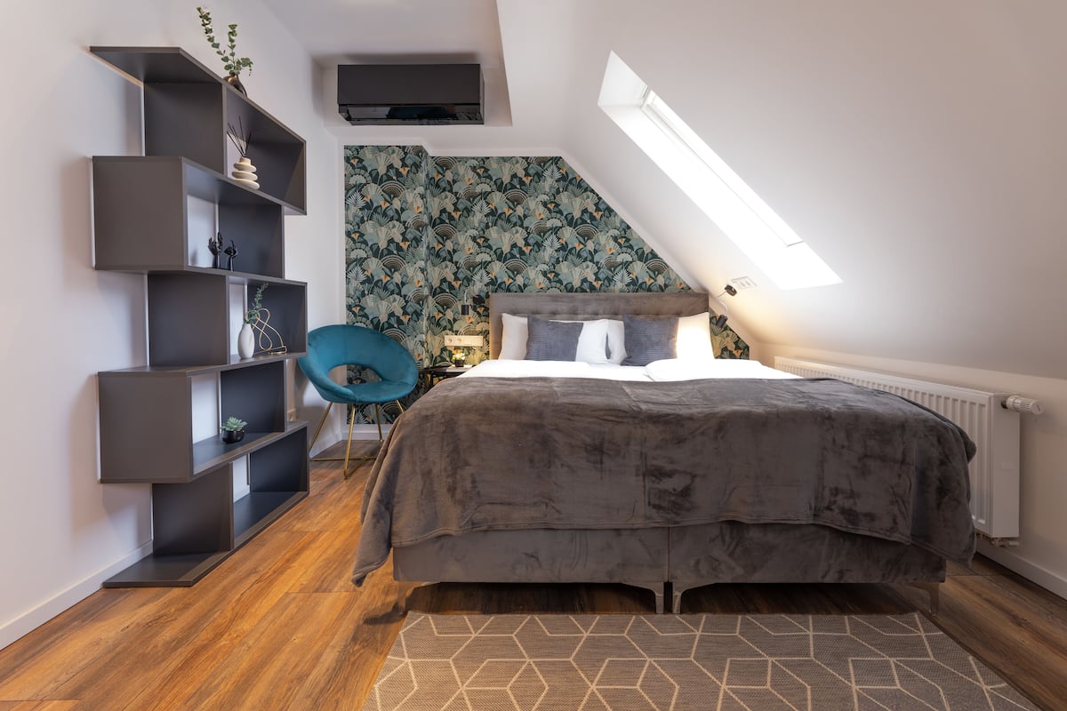 A bedroom with a sloped ceiling, a bed with a grey duvet, a blue chair, and a wall-mounted TV.
