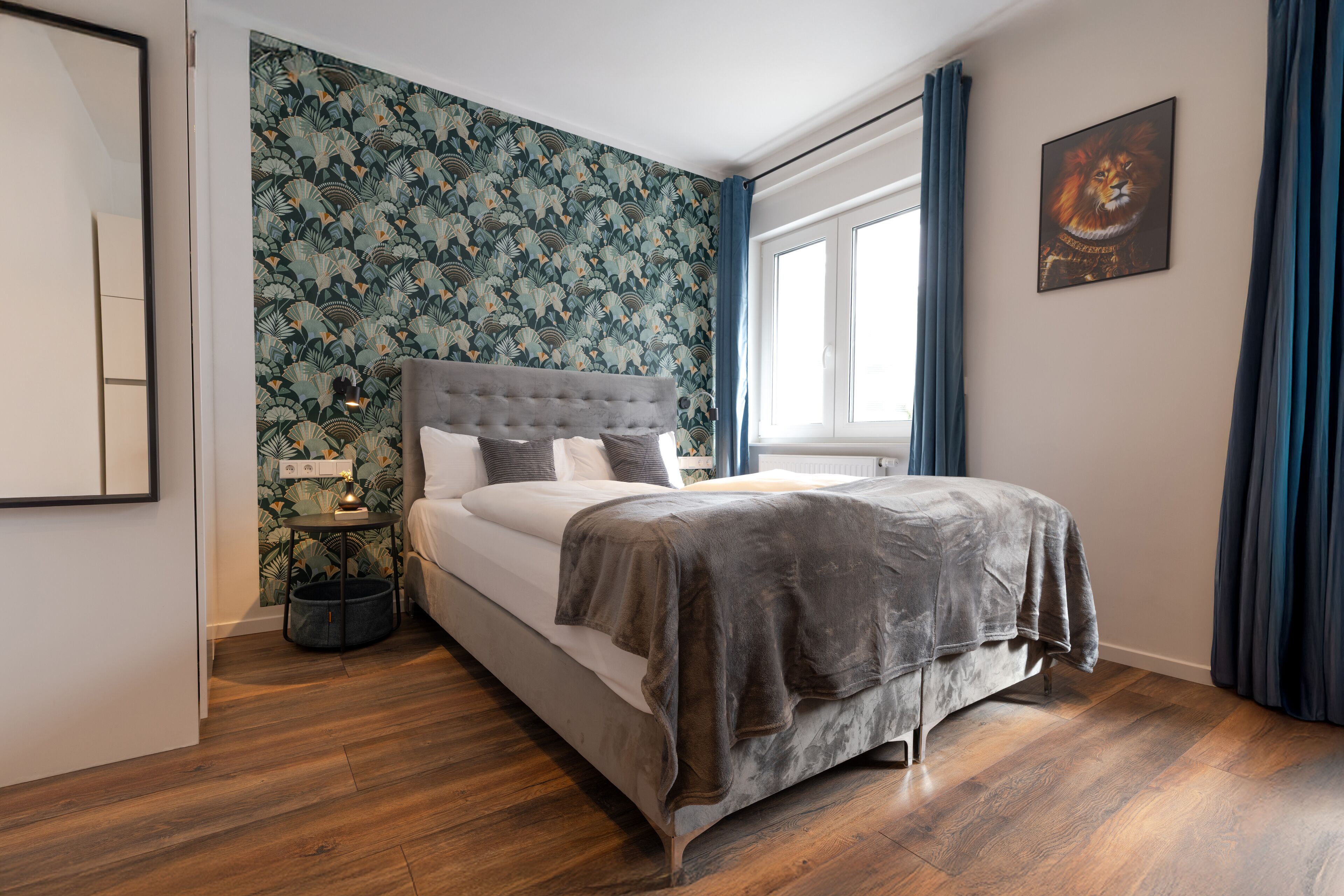 A bedroom with a bed, a nightstand, a wall with a floral wallpaper, a window with curtains, and a painting on the wall.